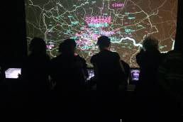 An image of four people looking at touch screens in front of a large map of London covered in words in pink and blue