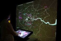 Image of a woman using a touch screen and looking at a large scale map of London with pink and blue words on it within the London Data Streams installation by Tekja